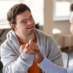 Young happy man with Down syndrome with his mentoring friend celebrating success indoors at school.