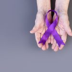 Hands of elderly woman hold purple ribbon on gray background. Alzheimer’s awareness concept.