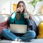 Cold woman working on laptop while drinking hot tea sitting on couch at home