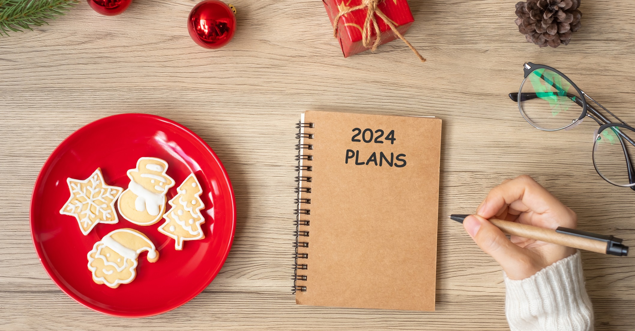 Woman hand writing 2024 PLANS on notebook with Christmas cookies on table. Xmas, Happy New Year, Goals, Resolution, To do list, and Strategy concept