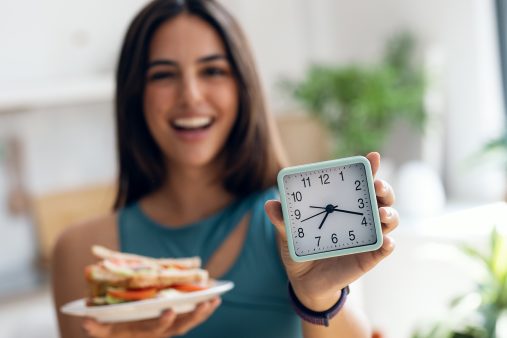 Happy sporty woman waiting for the time to eat while holding clock after fasting in the kitchen at home