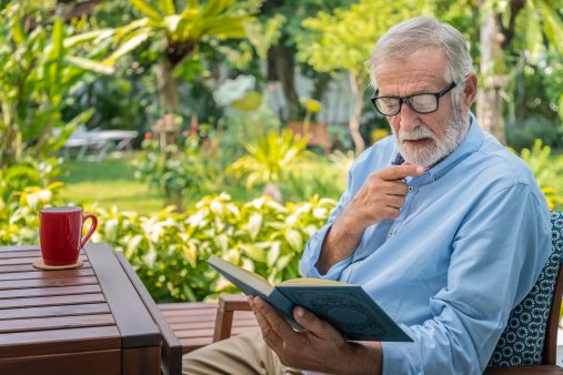 Senior elderly man reading book with mug of coffee in garden