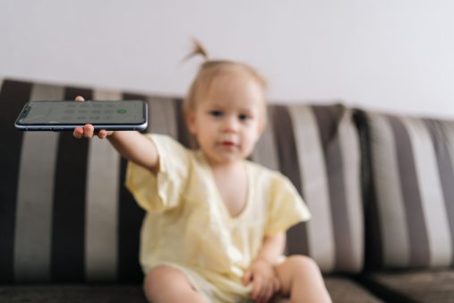 Fatiga digital: cómo afecta el exceso de pantallas al cerebro de adultos, niños y bebés Toddler girl looking at camera sitting on sofa, holding smartphone with cracked screen showing it forward.